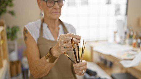 Foto de Dedicado senior artista de pelo gris, sosteniendo pinceles, profundamente absorto en la pintura en el estudio de arte. - Imagen libre de derechos
