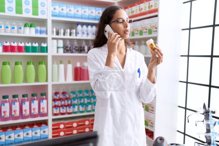 Foto de Young african american woman pharmacist holding pills bottle talking on smartphone at pharmacy - Imagen libre de derechos