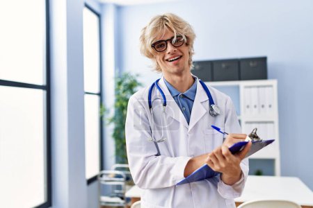 Photo for Young blond man doctor smiling confident writing on document at clinic - Royalty Free Image