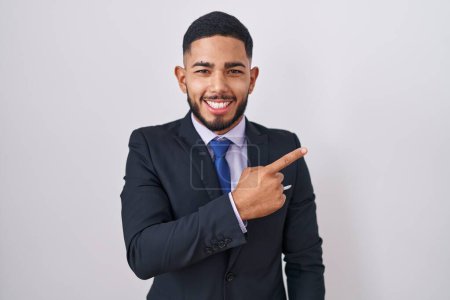 Foto de Joven hombre hispano con traje de negocios y corbata alegre con una sonrisa en la cara apuntando con la mano y el dedo hacia un lado con expresión feliz y natural - Imagen libre de derechos