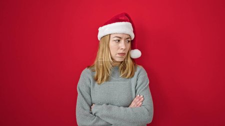 Photo for Young blonde woman wearing christmas hat standing with arms crossed gesture looking upset over isolated red background - Royalty Free Image