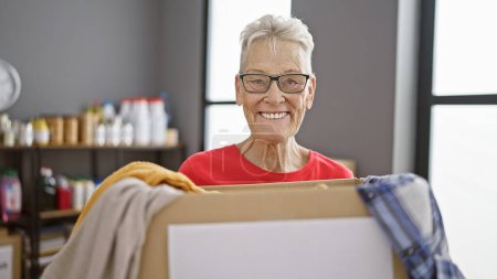 Téléchargez les photos : Une bénévole âgée aux cheveux gris confiante, souriante de façon générale, aime tenir un paquet de vêtements pour don au centre communautaire de bienfaisance. - en image libre de droit