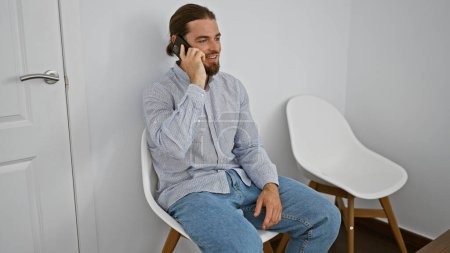 Téléchargez les photos : Jeune homme hispanique parlant sur smartphone assis sur une chaise souriant à la salle d'attente - en image libre de droit