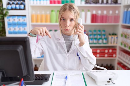 Photo for Young caucasian woman working at pharmacy drugstore speaking on the telephone pointing down looking sad and upset, indicating direction with fingers, unhappy and depressed. - Royalty Free Image
