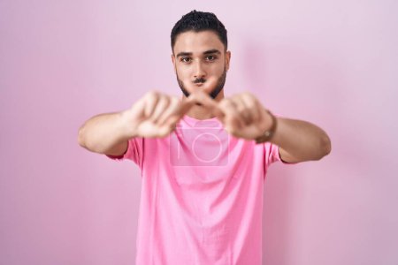 Foto de Joven hispano de pie sobre fondo rosa rechazo expresión cruzando dedos haciendo signo negativo - Imagen libre de derechos