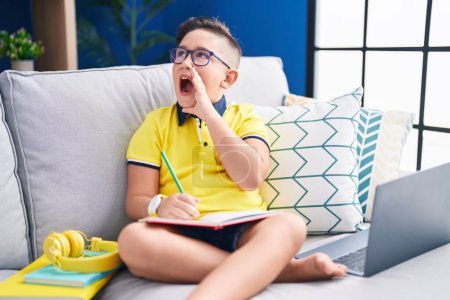 Foto de Joven niño hispano haciendo deberes sentado en el sofá gritando y gritando fuerte a un lado con la mano en la boca. concepto de comunicación. - Imagen libre de derechos