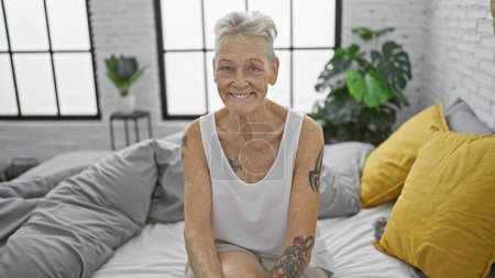 Téléchargez les photos : Femme âgée aux cheveux gris confiante savourant son matin, assise confortablement sur son lit confortable dans la chambre, son sourire vibrant reflétant un mode de vie mature. - en image libre de droit