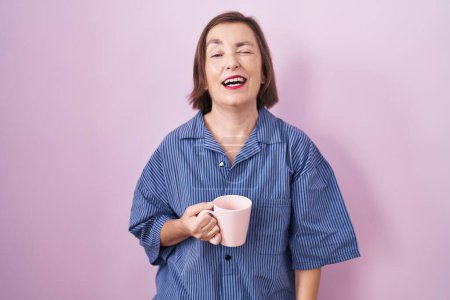 Téléchargez les photos : Moyen Age femme hispanique boire une tasse café clin d'oeil en regardant l'appareil photo avec une expression sexy, gai et heureux visage. - en image libre de droit