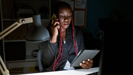 Photo for Concentrated african american woman boss talking business on smartphone, working on touchpad in office interior - Royalty Free Image