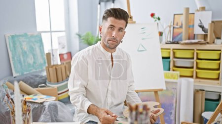 Foto de Atractivo joven hispano, un artista apasionado, concentrado en la pintura, sosteniendo el pincel y la paleta dentro de la animada sala de estudio de arte, provocando creatividad - Imagen libre de derechos