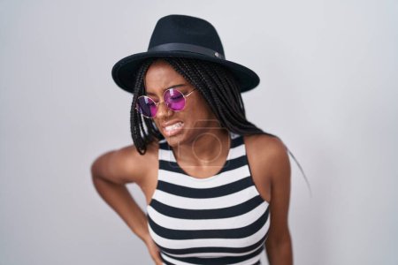 Téléchargez les photos : Jeune afro-américain avec des tresses portant chapeau et lunettes de soleil souffrant de maux de dos, toucher le dos avec la main, douleurs musculaires - en image libre de droit