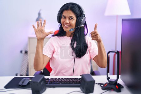 Foto de Mujer hispana madura jugando videojuegos en casa mostrando y señalando con los dedos número seis mientras sonríe confiada y feliz. - Imagen libre de derechos