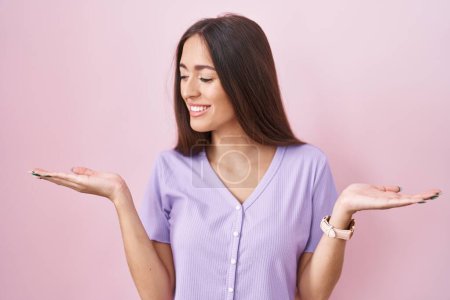 Photo for Young hispanic woman with long hair standing over pink background smiling showing both hands open palms, presenting and advertising comparison and balance - Royalty Free Image