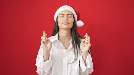 Téléchargez les photos : Jeune belle femme hispanique portant un chapeau de Noël croisant les doigts demandant de la chance sur fond rouge isolé - en image libre de droit
