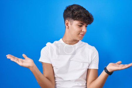 Téléchargez les photos : Adolescent hispanique debout sur fond bleu souriant montrant les paumes ouvertes des deux mains, présentant et comparant la publicité et l'équilibre - en image libre de droit