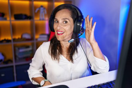 Téléchargez les photos : Femme brune du moyen âge jouant à des jeux vidéo souriant avec la main sur l'oreille écoutant et entendant des rumeurs ou des commérages. concept de surdité. - en image libre de droit