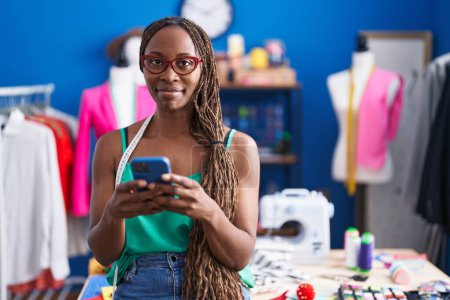 Téléchargez les photos : Femme afro-américaine tailleur souriant confiant en utilisant smartphone à l'atelier - en image libre de droit