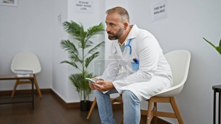 Photo for Handsome young man, a serious-faced doctor, texting on smartphone while relaxedly sitting in clinic's waiting room chair immersed in his medical work. - Royalty Free Image