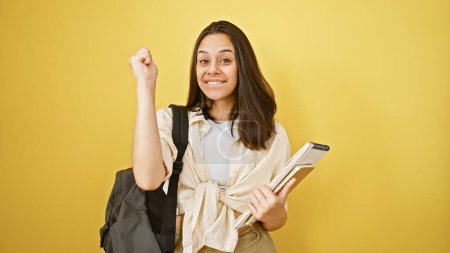 Foto de Retrato festivo de una joven y hermosa estudiante hispana confiada sosteniendo libros, de pie aislada sobre un vibrante fondo amarillo, exudando una sonrisa ganadora y un estilo de vida casual - Imagen libre de derechos