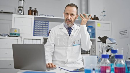 Foto de Hombre hispano de mediana edad de cabello gris, un científico de laboratorio, absorto en la investigación científica, escuchando un mensaje de voz en su teléfono inteligente en la mesa del laboratorio - Imagen libre de derechos