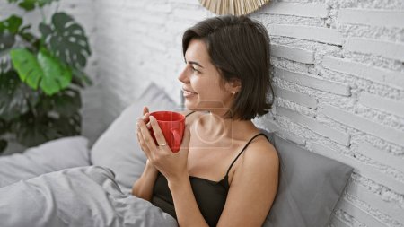 Foto de Mujer hispana joven relajada sentada cómodamente en su cama, oliendo el aroma encantador del café de la mañana. hermosa mujer en su dormitorio disfrutando de su bebida con una sonrisa segura. - Imagen libre de derechos