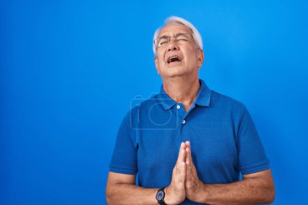 Téléchargez les photos : Homme du Moyen Âge avec les cheveux gris debout sur fond bleu mendier et prier avec les mains ainsi que l'expression de l'espoir sur le visage très émotionnel et inquiet. mendier. - en image libre de droit