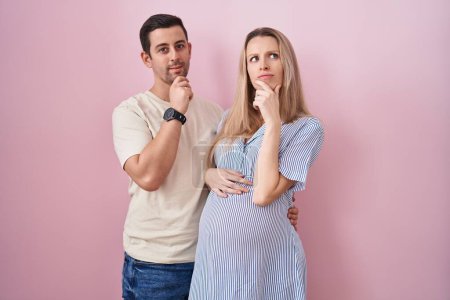 Photo for Young couple expecting a baby standing over pink background looking confident at the camera smiling with crossed arms and hand raised on chin. thinking positive. - Royalty Free Image