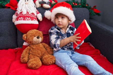 Foto de Adorable niño hispano leyendo libro sentado en el sofá por la decoración de Navidad en casa - Imagen libre de derechos