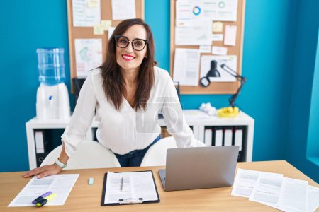 Foto de Young beautiful hispanic woman business worker smiling confident standing by desk at office - Imagen libre de derechos