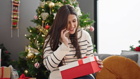 Photo for Young beautiful hispanic woman holding christmas gift talking on smartphone at home - Royalty Free Image
