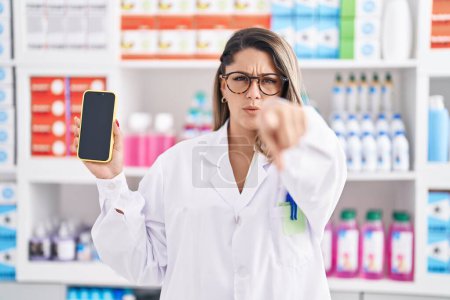 Foto de Mujer rubia que trabaja en la farmacia mostrando la pantalla del teléfono inteligente señalando con el dedo a la cámara y a usted, gesto de confianza que se ve serio - Imagen libre de derechos