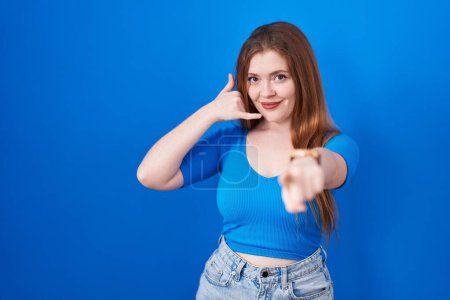 Téléchargez les photos : Femme rousse debout sur fond bleu souriant faire parler au téléphone geste et vous pointant du doigt. Appelle-moi. - en image libre de droit