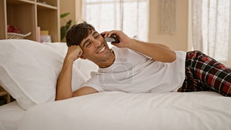 Téléchargez les photos : Heureux, confiant jeune homme hispanique profiter d'une conversation matinale relaxante sur son téléphone, couché confortablement sur son lit dans la chambre de son appartement. - en image libre de droit