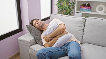 Foto de Hombre hispano joven preocupado, retrato de la enfermedad, sufriendo dolor de estómago intenso mientras descansa infelizmente, acostado en el sofá de la sala de estar, en el interior, en casa - Imagen libre de derechos