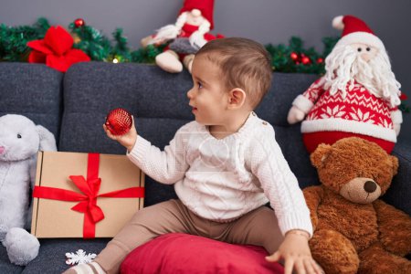 Photo for Adorable hispanic toddler holding christmas ball sitting on sofa at home - Royalty Free Image
