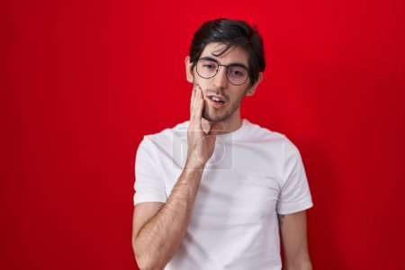 Téléchargez les photos : Jeune homme hispanique debout sur fond rouge touchant la bouche avec la main avec une expression douloureuse à cause de maux de dents ou de maladies dentaires sur les dents. dentiste - en image libre de droit