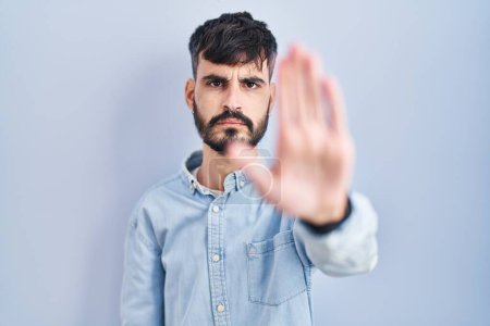 Photo for Young hispanic man with beard standing over blue background doing stop sing with palm of the hand. warning expression with negative and serious gesture on the face. - Royalty Free Image