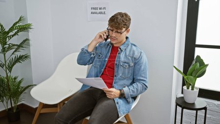 Photo for Handsome young hispanic man engaged in a digital conversation over his mobile phone, reading important paperwork while comfortably sitting on a chair within the indoors of a waiting room - Royalty Free Image