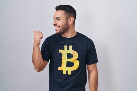 Téléchargez les photos : Jeune homme hispanique portant Bitcoin t-shirt souriant avec le visage heureux regardant et pointant vers le côté avec le pouce vers le haut. - en image libre de droit