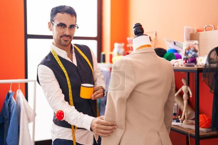 Téléchargez les photos : Jeune homme hispanique tailleur boire du café debout par mannequin à l'atelier - en image libre de droit