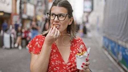 Photo for Stunning hispanic woman savors sweet potato snack in her eyeglasses on the buzzing osaka's dotonbori street - Royalty Free Image