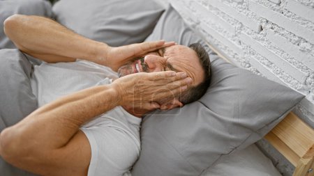 Téléchargez les photos : Homme d'âge moyen avec les cheveux gris couché sur le lit souffrant de maux de tête dans la chambre - en image libre de droit