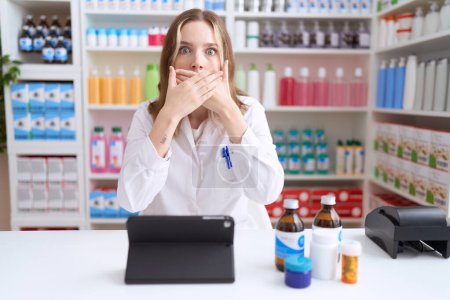 Téléchargez les photos : Jeune femme caucasienne travaillant à la pharmacie pharmacie en utilisant comprimé choqué couvrant la bouche avec les mains pour erreur. concept secret. - en image libre de droit