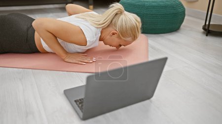Photo for Sporty, attractive and fit young blonde woman staying healthy - stretching back in calm balance during an online yoga class in her beautiful living room - Royalty Free Image