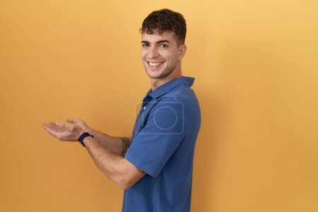 Téléchargez les photos : Jeune homme hispanique debout sur fond jaune pointant de côté avec les mains ouvertes paumes montrant l'espace de copie, présentant la publicité souriant heureux excité - en image libre de droit