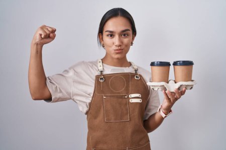 Téléchargez les photos : Jeune femme hispanique portant tablier de serveuse professionnel tenant café personne forte montrant muscle du bras, confiant et fier de la puissance - en image libre de droit
