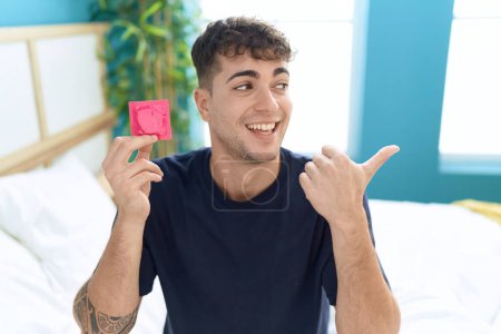 Téléchargez les photos : Jeune homme hispanique assis sur le lit tenant préservatif pointant le pouce vers le côté souriant heureux avec la bouche ouverte - en image libre de droit