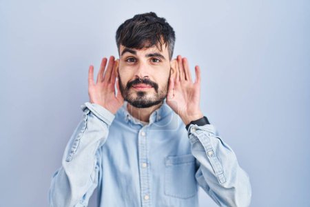 Foto de Joven hombre hispano con barba de pie sobre fondo azul tratando de escuchar ambas manos en el gesto de la oreja, curioso por chismes. problema auditivo, sordo - Imagen libre de derechos