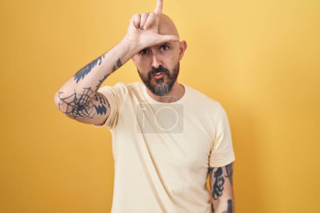 Photo for Hispanic man with tattoos standing over yellow background making fun of people with fingers on forehead doing loser gesture mocking and insulting. - Royalty Free Image