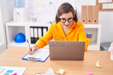 Photo for Middle age woman call center agent writing on document working at office - Royalty Free Image